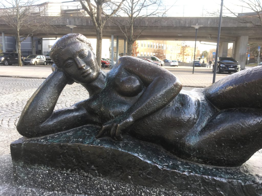 Eric Grates skulptur "Liggande kvinna" vilar vid kanten av fontändammen i Västertorps centrum. Bakom henne går tunnelbanetågen.