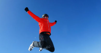 En man i röd skidjacka och hjälm hoppar med armarna utsträcka, och mot bakgrund av en klarblå himmel.