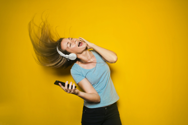 Dansande tjej med hörlurar och mobilen i handen. Stark gul bakgrund och långt hår som flyger.